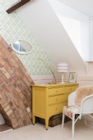 Yellow painted chest of drawers in the corner of an attic bedroom with exposed brick chimney breast