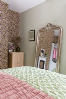 A large ornate gold framed mirror at the foot of a bed leans against a pale green wall in a bedroom with a feature wall papered with a leopard print design.