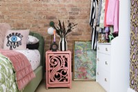 An exposed brick wall in a bedroom with a pink painted rattan bedside table next to a green velvet bed and an open grab and go wardrobe and drawer unit.