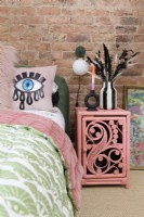 An exposed brick wall in a bedroom with a pink painted rattan bedside table next to a green velvet bed