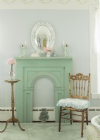 A convex mirror hangs above a fireplace surround painted in distressed green. A gilded chair and table instill a touch of faded glamor.