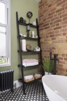 Black ladder shelving unit leaning against a green wall in the corner of a modern bathroom with an exposed brick wall