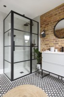 A crittall shower enclosure in the corner of a modern bathroom with an exposed brick wall and a monochrome patterned tiled floor
