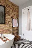 A black towel rail in the corner of a bathroom with exposed brickwork and a white door against a smooth grey cement wall with a monochrome patterned tiled floor