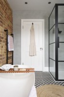 View along a bath tub to a white bathroom door and grey smooth cement wall with an exposed brick wall on one side and a crittall shower enclosure on the other side