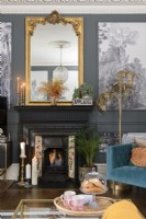 Black painted Victorian fireplace with gold mantle mirror in a grey painted living room with paneling filled with monochrome tropical tree wallpaper
