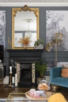 Black painted Victorian fireplace with gold mantle mirror in a grey painted living room with paneling filled with monochrome tropical tree wallpaper