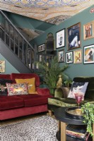 Open plan green living room with red and green velvet sofas in front of an open staircase and a salon style gallery art display wall and wall papered mosaic patterned ceiling
