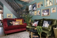 Open plan green living room with red and green velvet sofas in front of an open staircase and a salon style gallery art display wall
