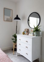 White chest of drawers and black framed round mirror in bedroom